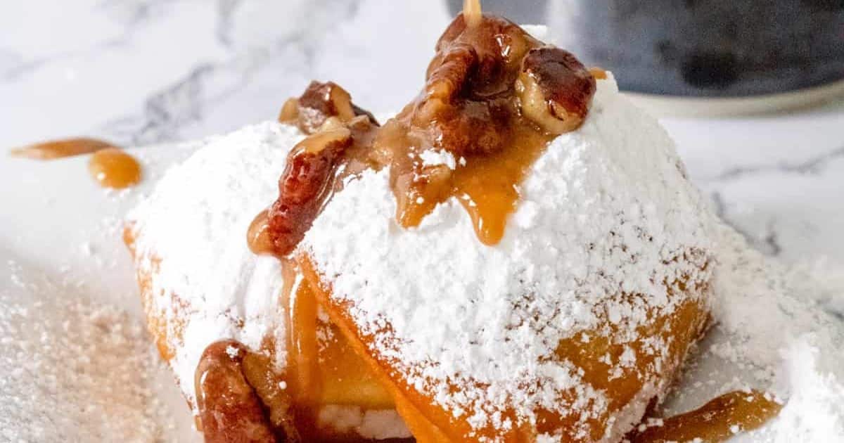 Beignet topped with powdered sugar, caramel sauce, and pecans on a white surface.