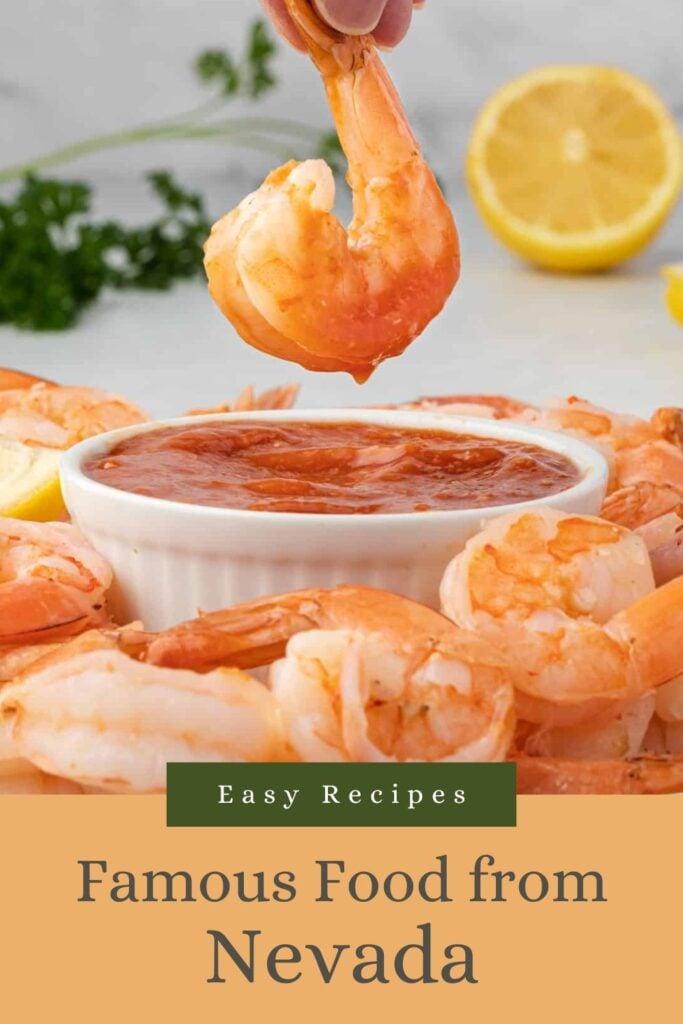 A hand dips a shrimp into cocktail sauce, with more shrimp, lemon, and parsley in the background.