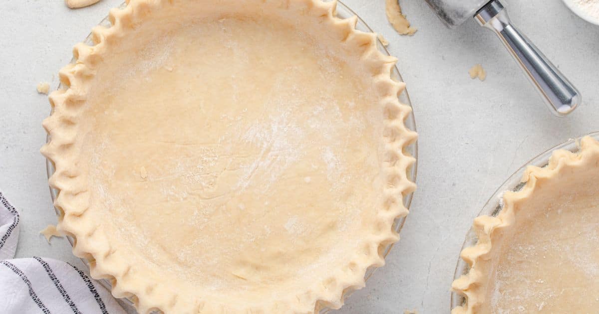 Unbaked pie crust with crimped edges in a glass dish on a light surface, with dough scraps nearby.