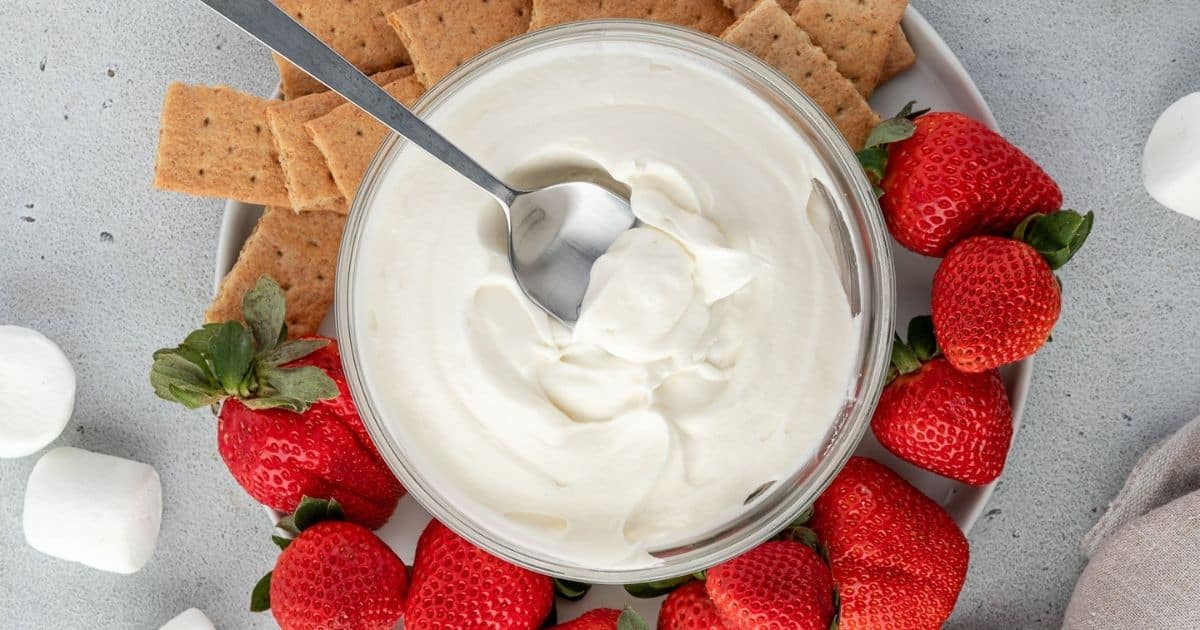 A bowl of creamy dip with a spoon, surrounded by strawberries, graham crackers, and marshmallows.