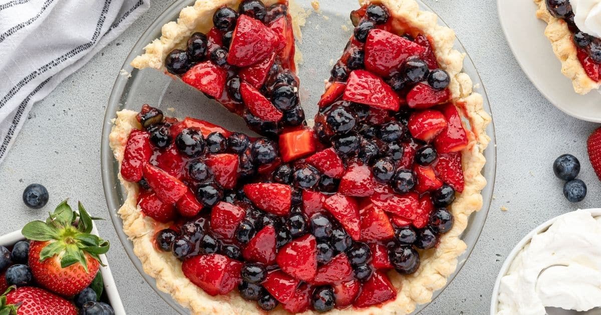 A berry pie with strawberries and blueberries, with two slices missing, on a light-colored table.