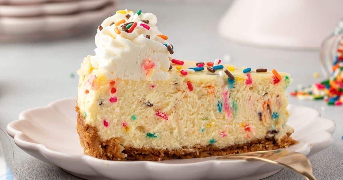 A slice of funfetti cheesecake with whipped cream and colorful sprinkles on a plate.
