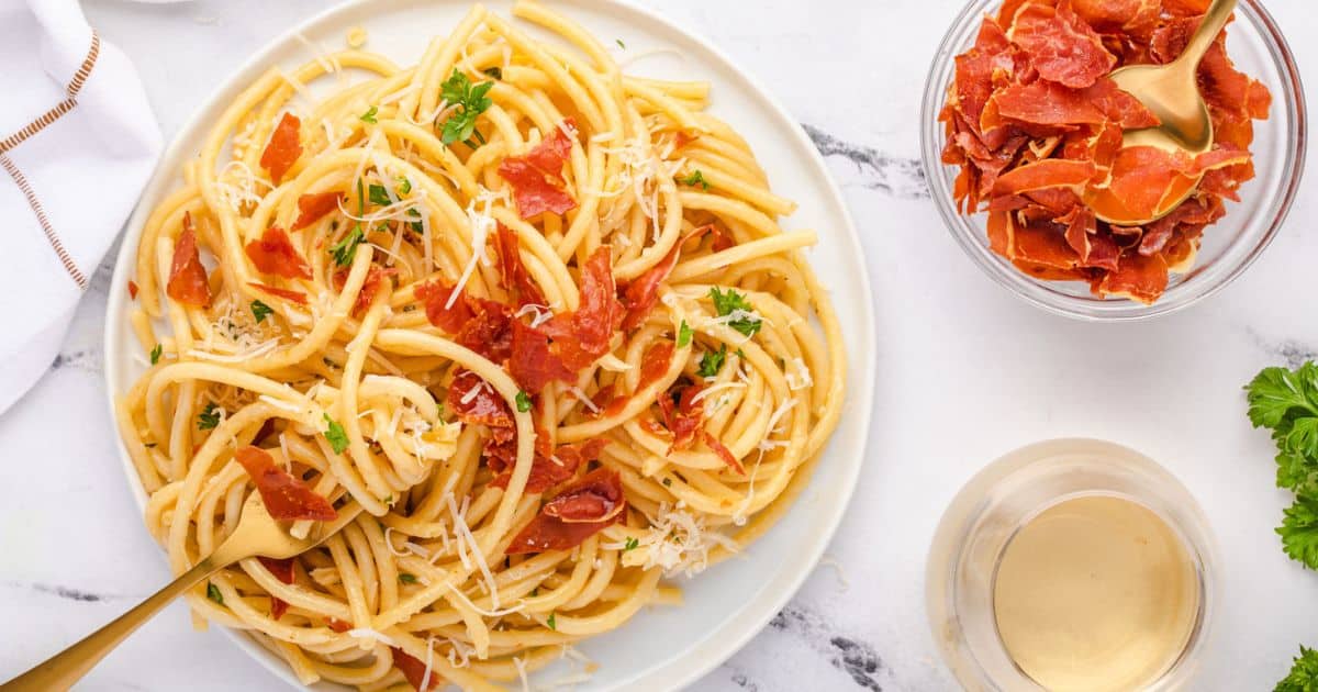 Plate of pasta with crispy prosciutto and grated cheese, next to a bowl of prosciutto and a glass of white wine.