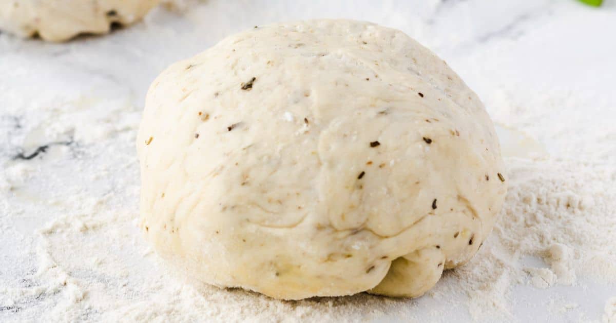 A ball of herbed pizza dough sits on a floured surface, ready to be rolled out.