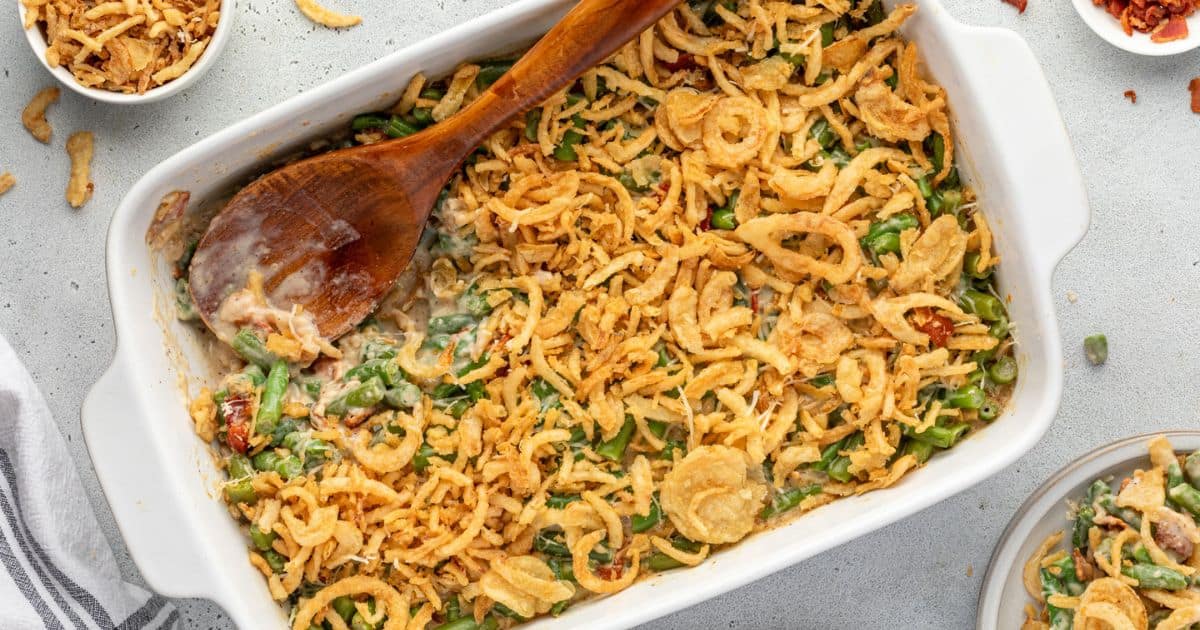 Green bean casserole topped with crispy fried onions in a white baking dish, with a wooden spoon inside.