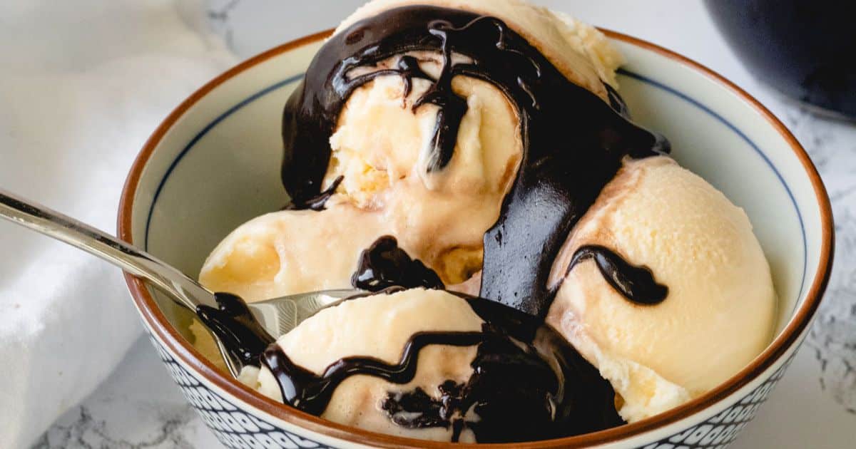Vanilla ice cream scoops in a bowl topped with chocolate syrup, with a spoon inside the bowl.