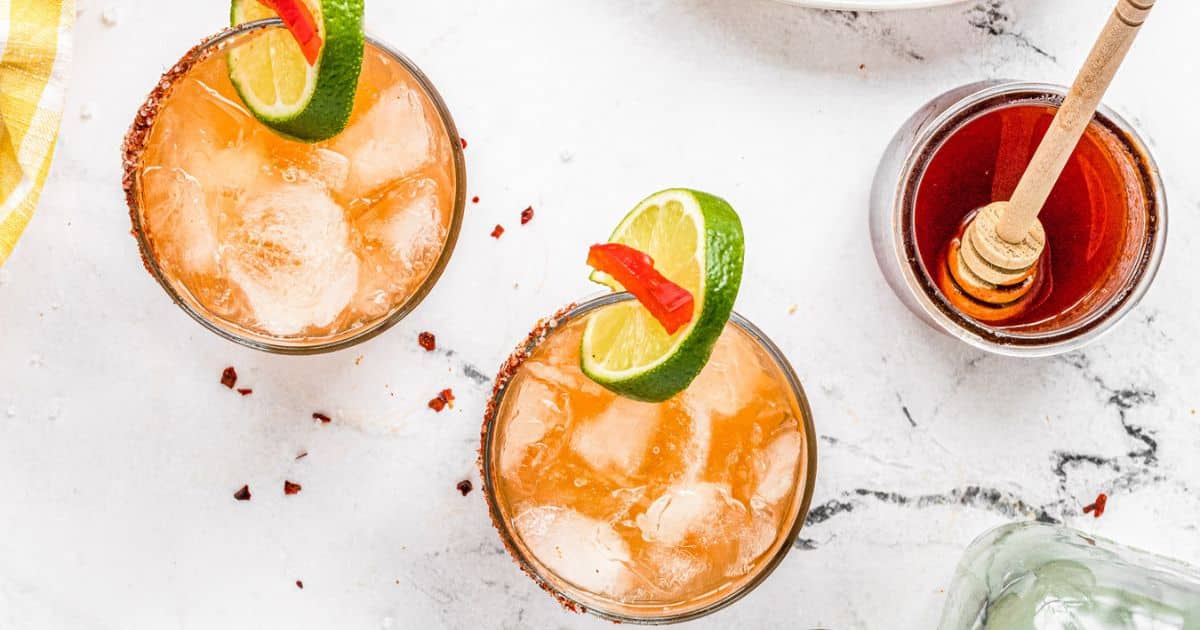 Two iced cocktails with lime and chili garnish, next to a jar of honey with a dipper on a white marble surface.