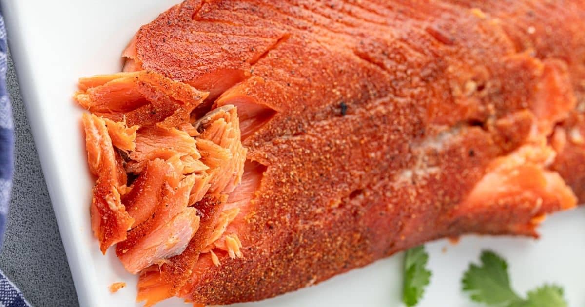 Close-up of a seasoned, flaky baked salmon fillet on a white plate with a sprig of cilantro nearby.