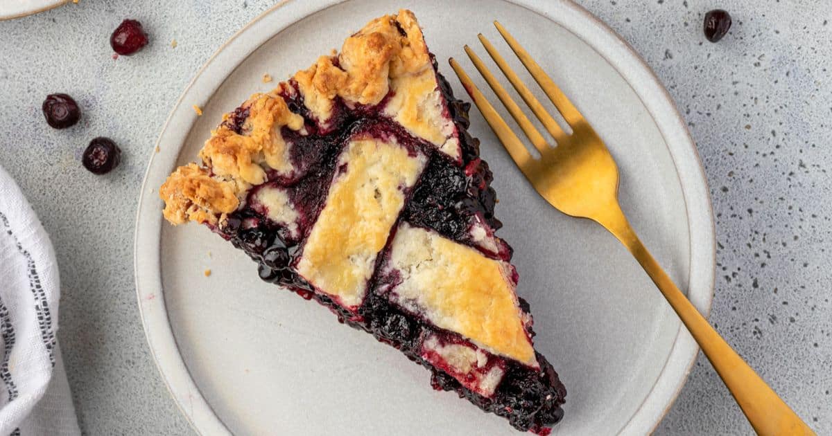 A slice of blueberry pie with a lattice crust on a plate next to a gold fork.