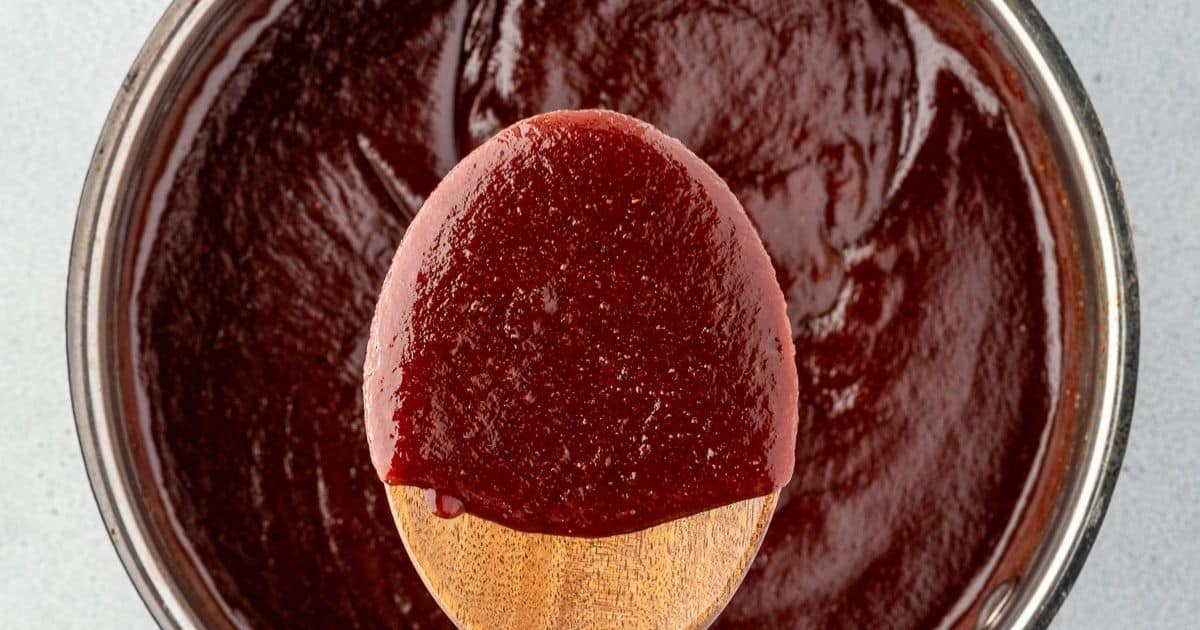 A wooden spoon dipped in thick, dark red barbecue sauce over a bowl of the same sauce.