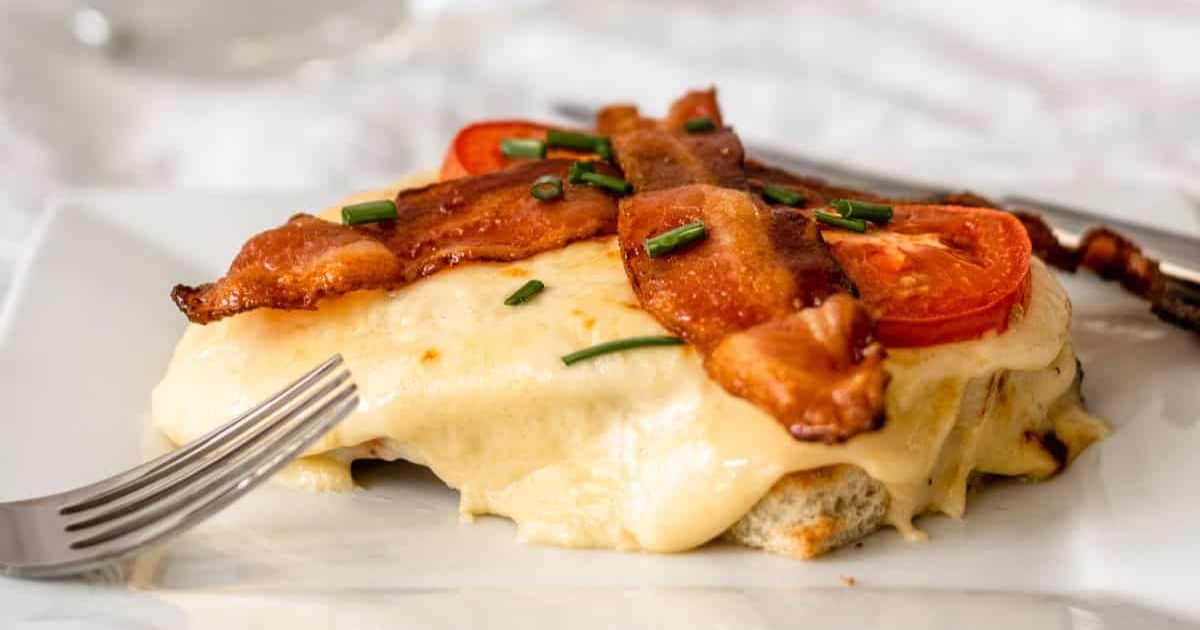 Open-faced sandwich with melted cheese, bacon, tomato slices, and chives on a white plate with a fork.