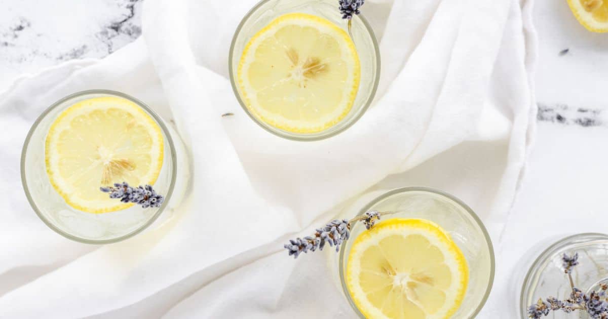 Glasses of lemonade with lemon slices and lavender sprigs on a white cloth background.