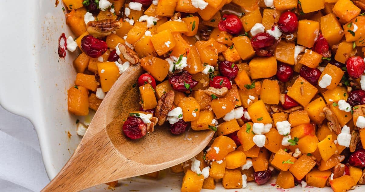 Roasted butternut squash with cranberries, pecans, and goat cheese in a white dish with a wooden spoon.