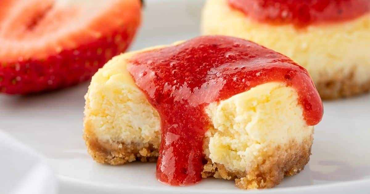 A mini cheesecake with a bite taken out, topped with strawberry sauce, next to a sliced strawberry.