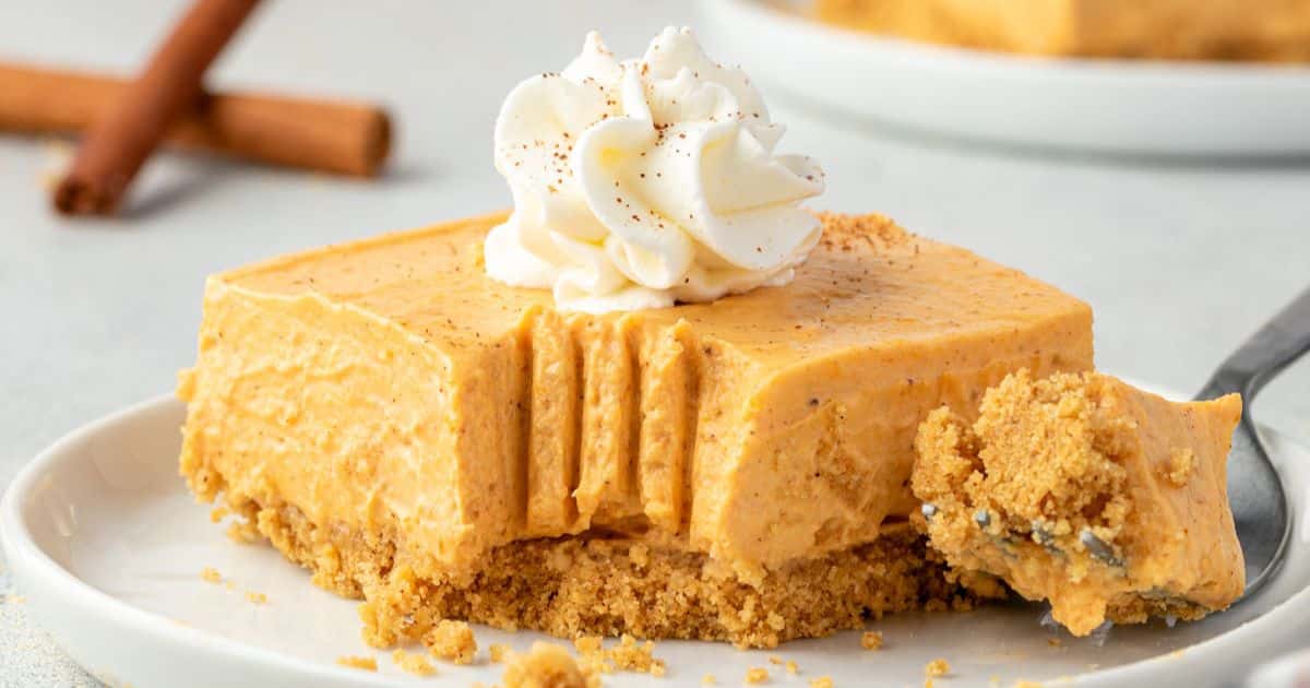 A slice of creamy pumpkin cheesecake with whipped cream on top and a bite taken out, on a white plate.