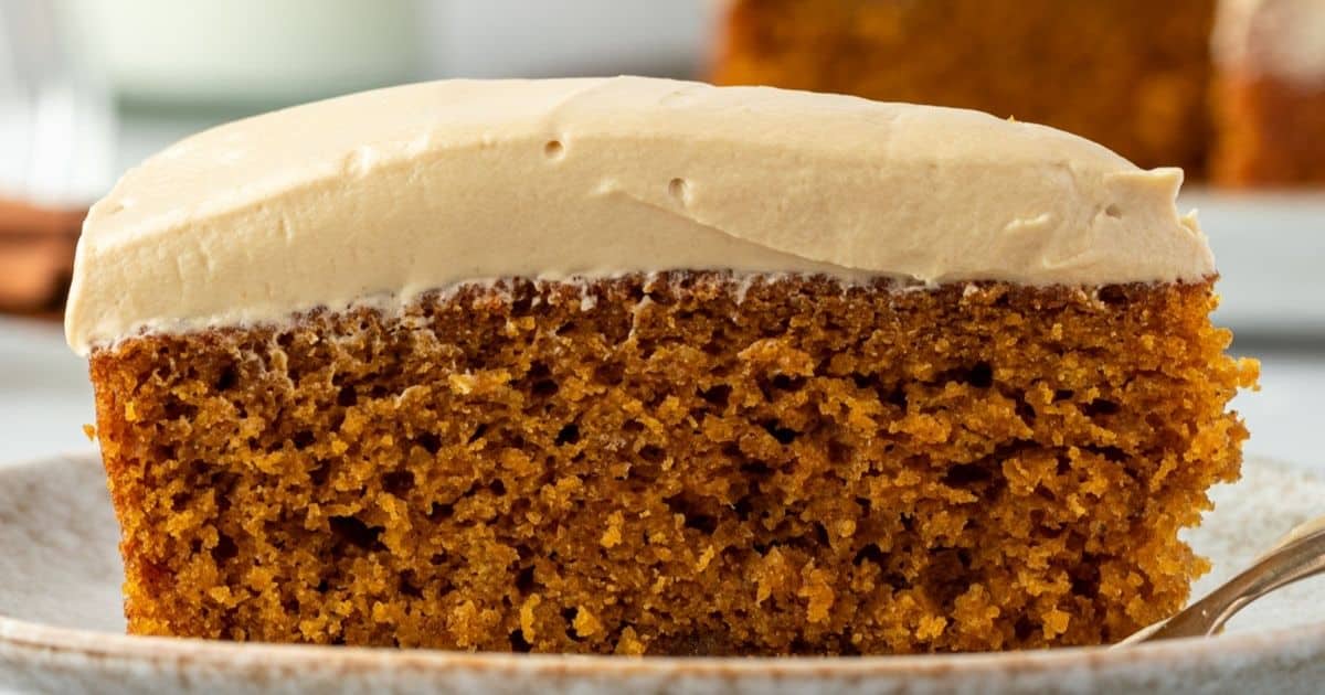 A slice of moist brown cake with thick, creamy frosting on top, served on a beige plate.