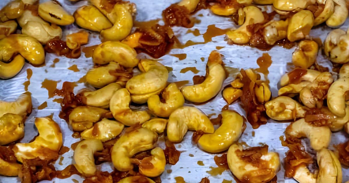 Cashew nuts coated in a reddish seasoning are spread out on a metallic surface.