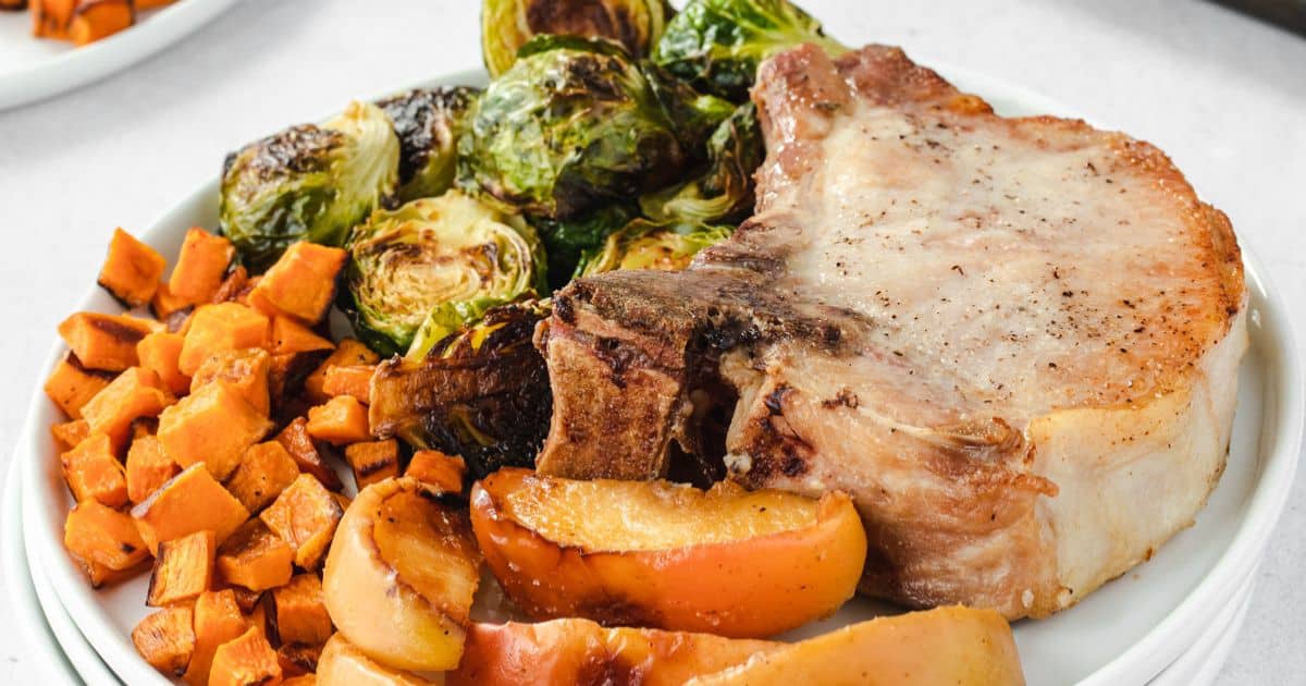 A plate with a pork chop, roasted brussels sprouts, diced sweet potatoes, and sliced roasted apples.