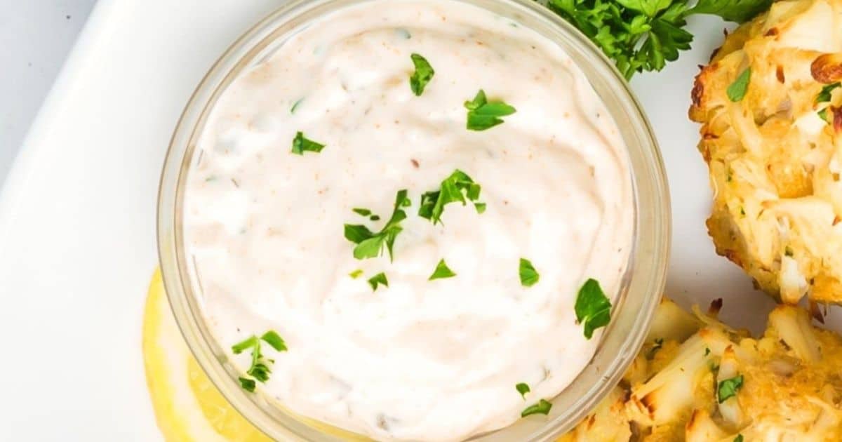 A bowl of creamy white dipping sauce garnished with chopped parsley, next to crab cakes and a lemon slice.