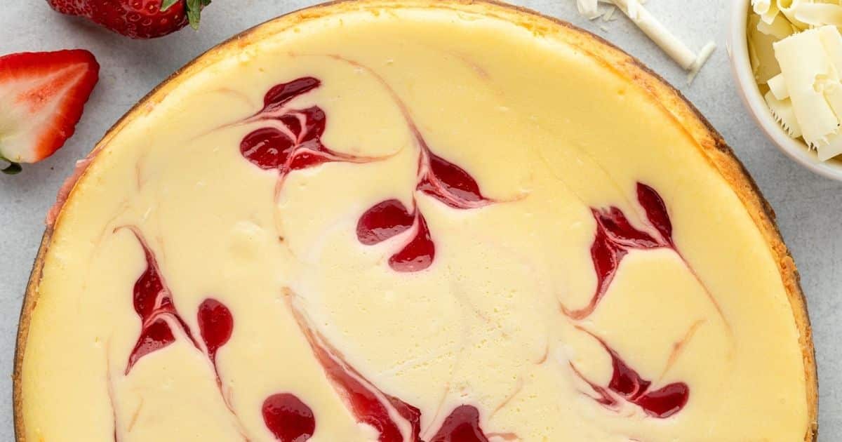 A cheesecake with red strawberry swirls on top, surrounded by strawberries and white chocolate curls.