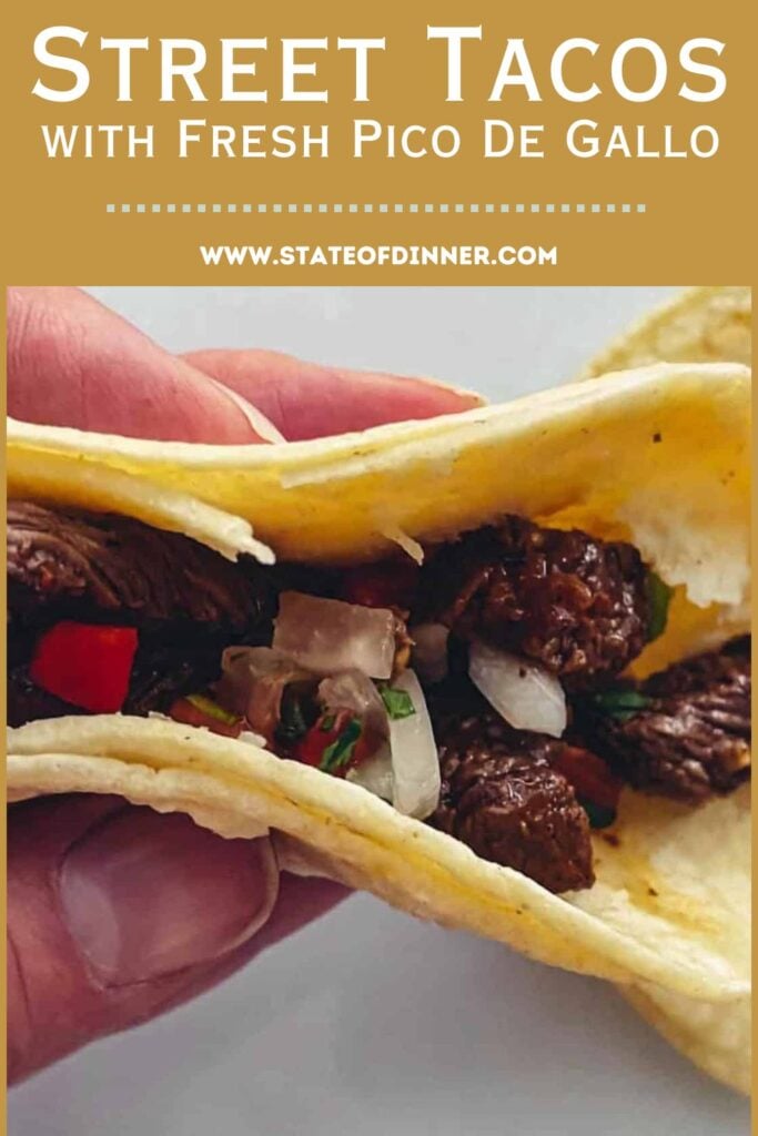 Hand holding a street taco filled with beef, onions, peppers, and fresh pico de gallo in a corn tortilla.