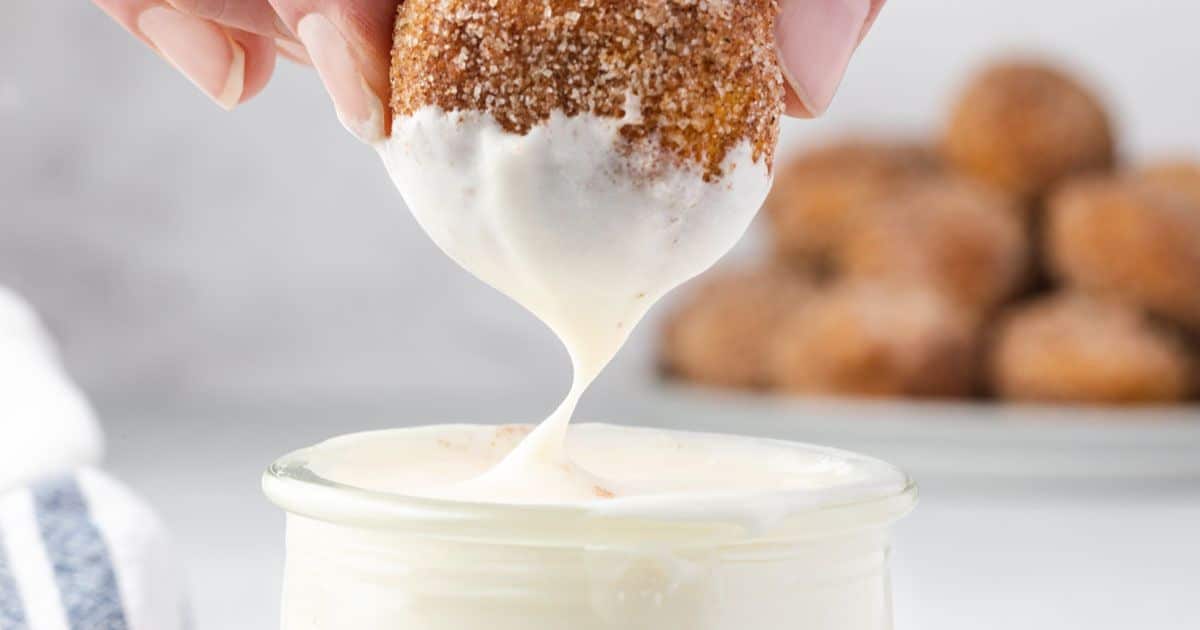 A hand dips a cinnamon-sugar donut hole into creamy white icing, with more donut holes in the background.