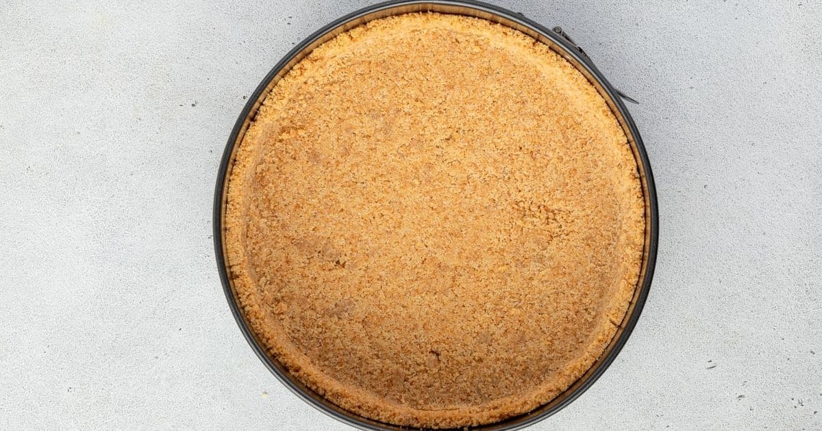 Round cheesecake crust in a springform pan on a light gray surface, viewed from above.