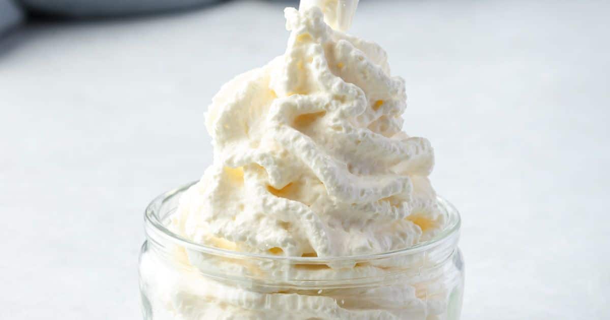 Whipped cream swirled high in a small, clear glass jar against a light background.