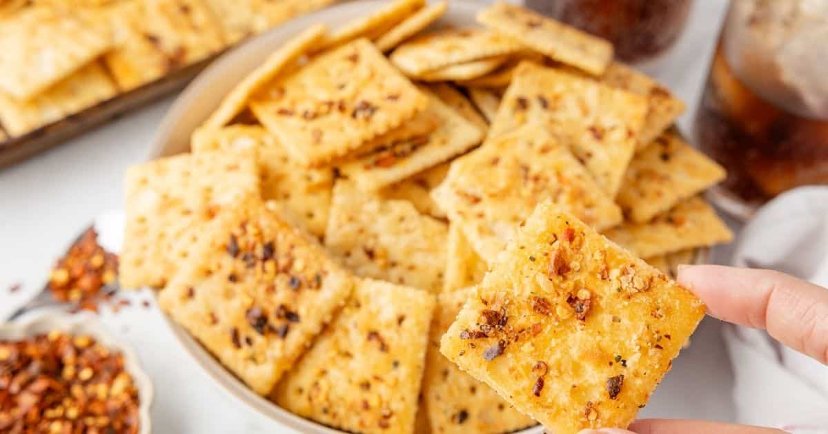 A hand holds a seasoned cracker above a bowl filled with spicy, seasoned crackers.