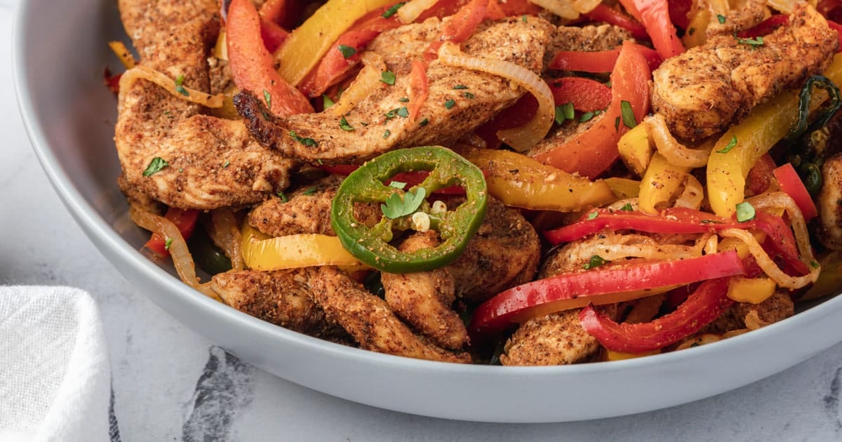 Sliced chicken with red and yellow bell peppers, onions, and jalapeño in a gray bowl.