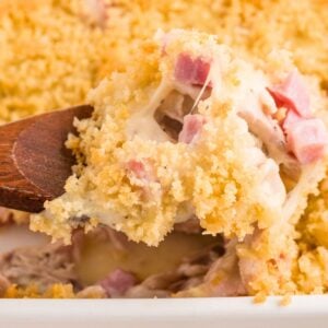 A wooden spoon scoops cheesy ham casserole with a golden breadcrumb topping from a baking dish.