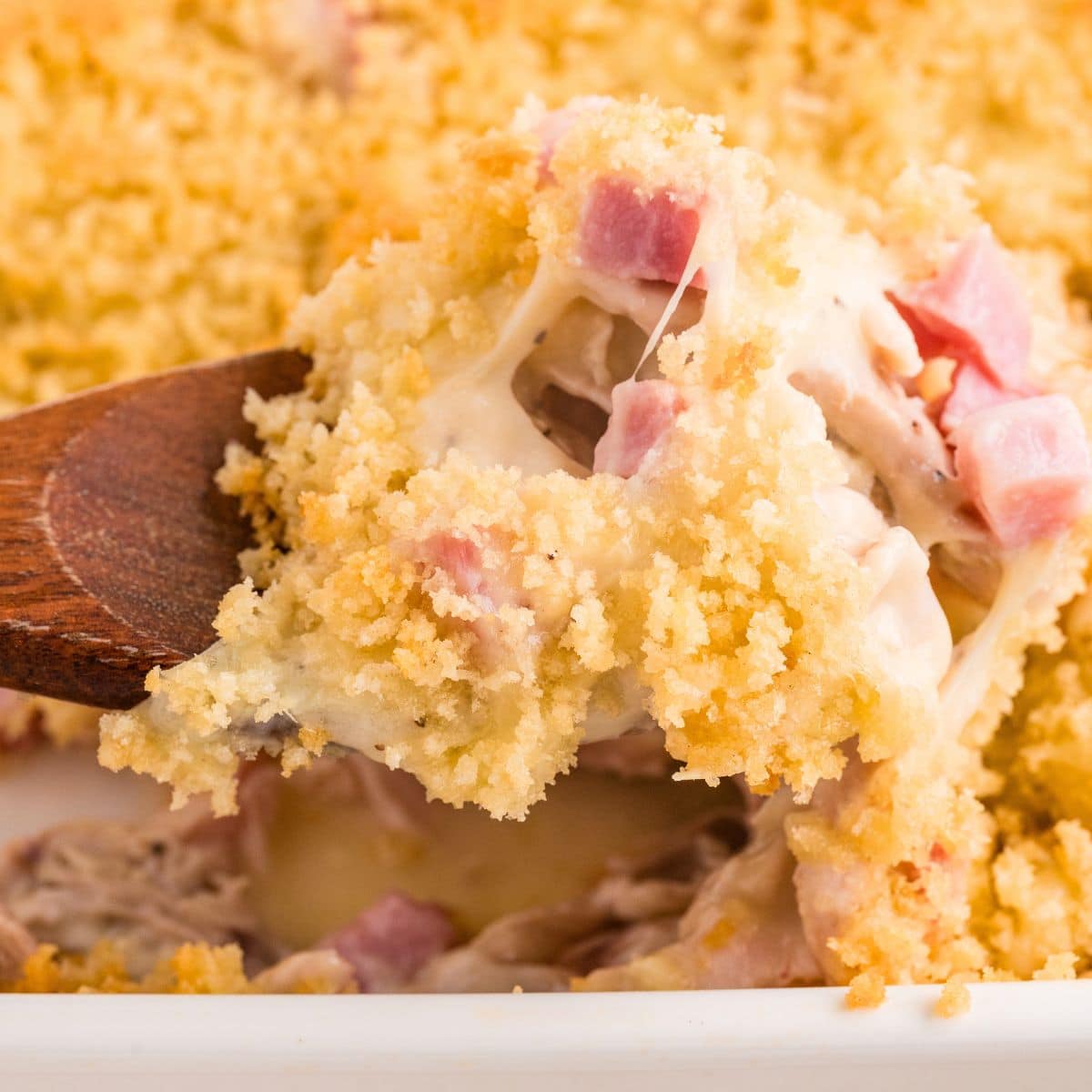 A wooden spoon scoops cheesy ham casserole with a crispy breadcrumb topping from a white baking dish