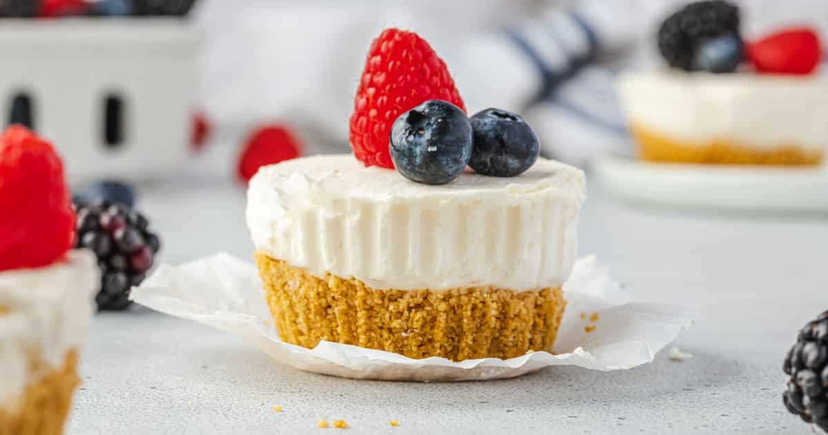 Mini cheesecake with a graham cracker crust, topped with a raspberry and blueberries, on a white surface.