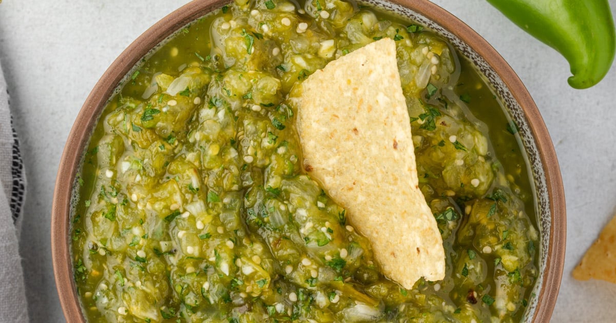 A bowl of green salsa with a tortilla chip dipped in it, photographed from above.