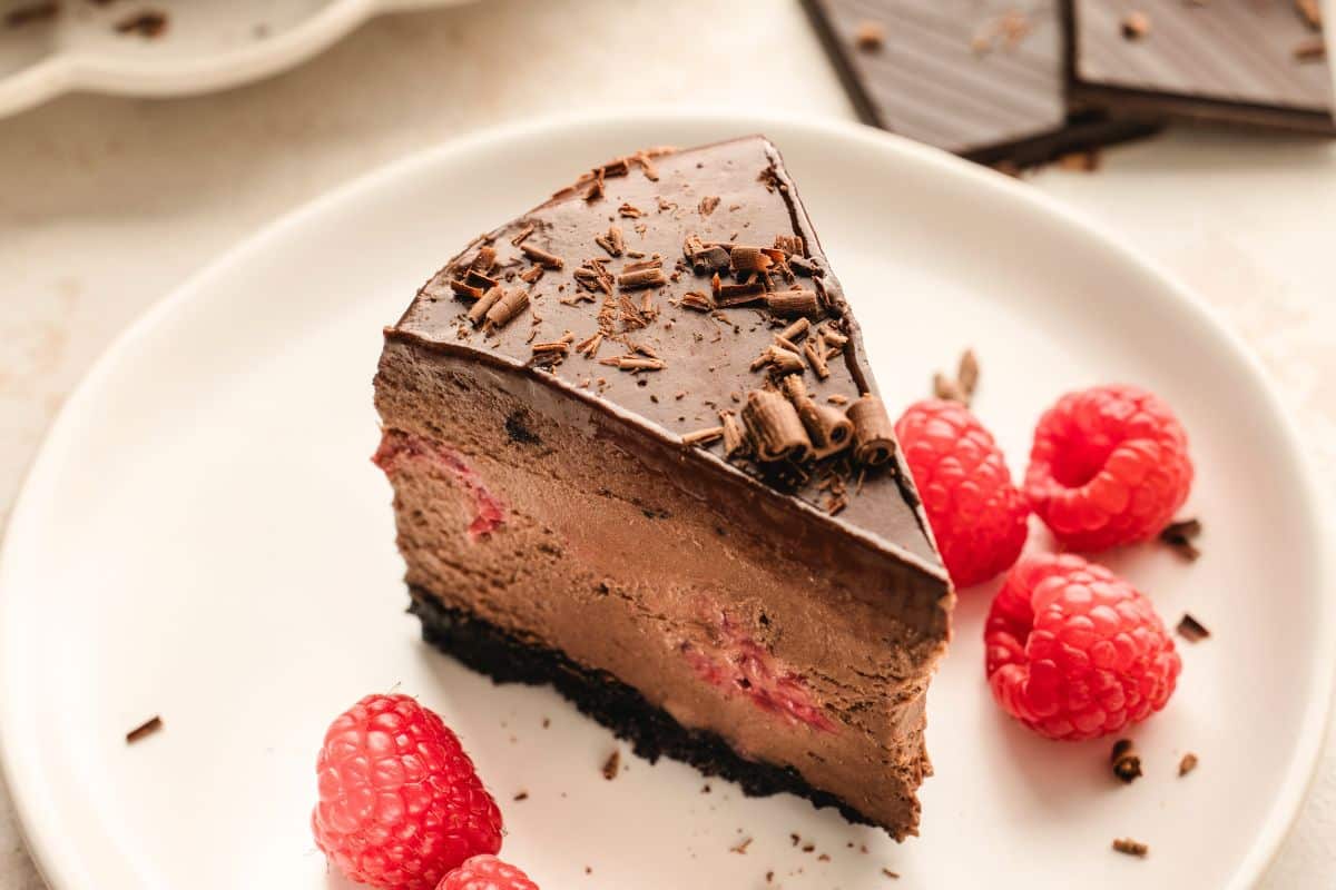 A slice of chocolate cheesecake with chocolate shavings and raspberries on a white plate.