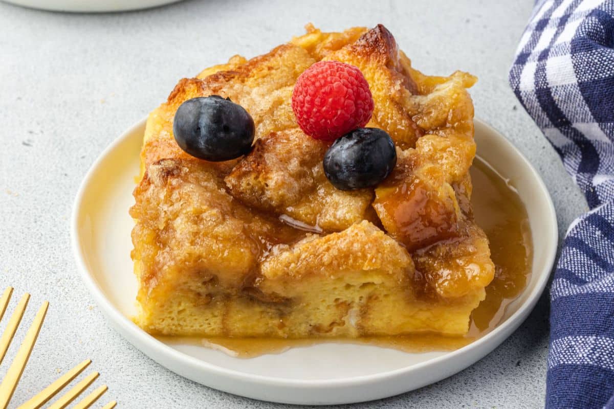 A slice of bread pudding topped with syrup, a raspberry, and two blueberries on a white plate.
