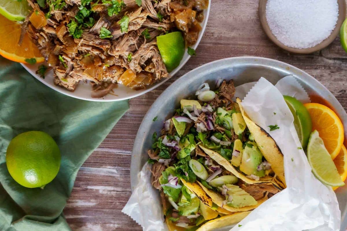 Three tacos with shredded meat, onions, cilantro, and avocado, next to a bowl of shredded meat and lime slices.