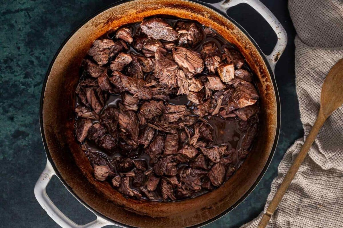 Chunks of braised beef in a Dutch oven, surrounded by a wooden spoon and a textured cloth.