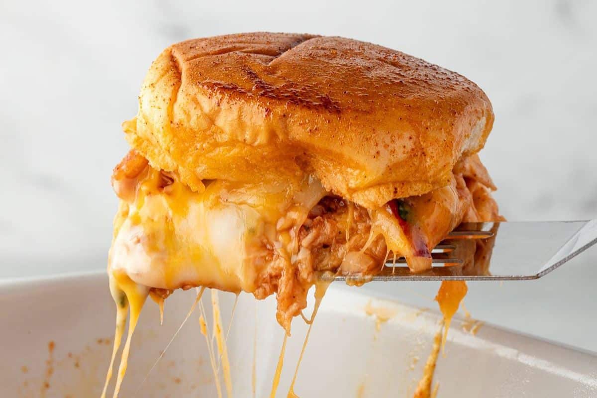 A close-up of a cheesy, pulled chicken sandwich on a spatula, with melted cheese stretching.