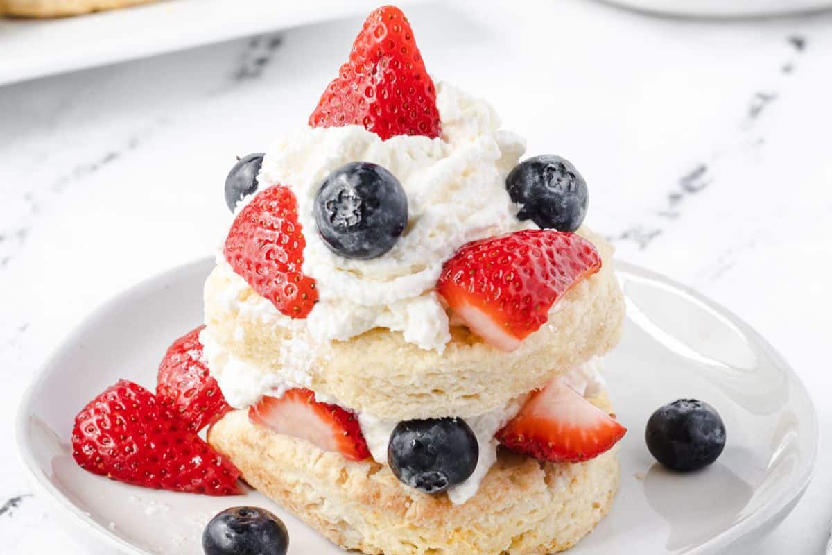 A biscuit topped with whipped cream, strawberries, and blueberries on a white plate.