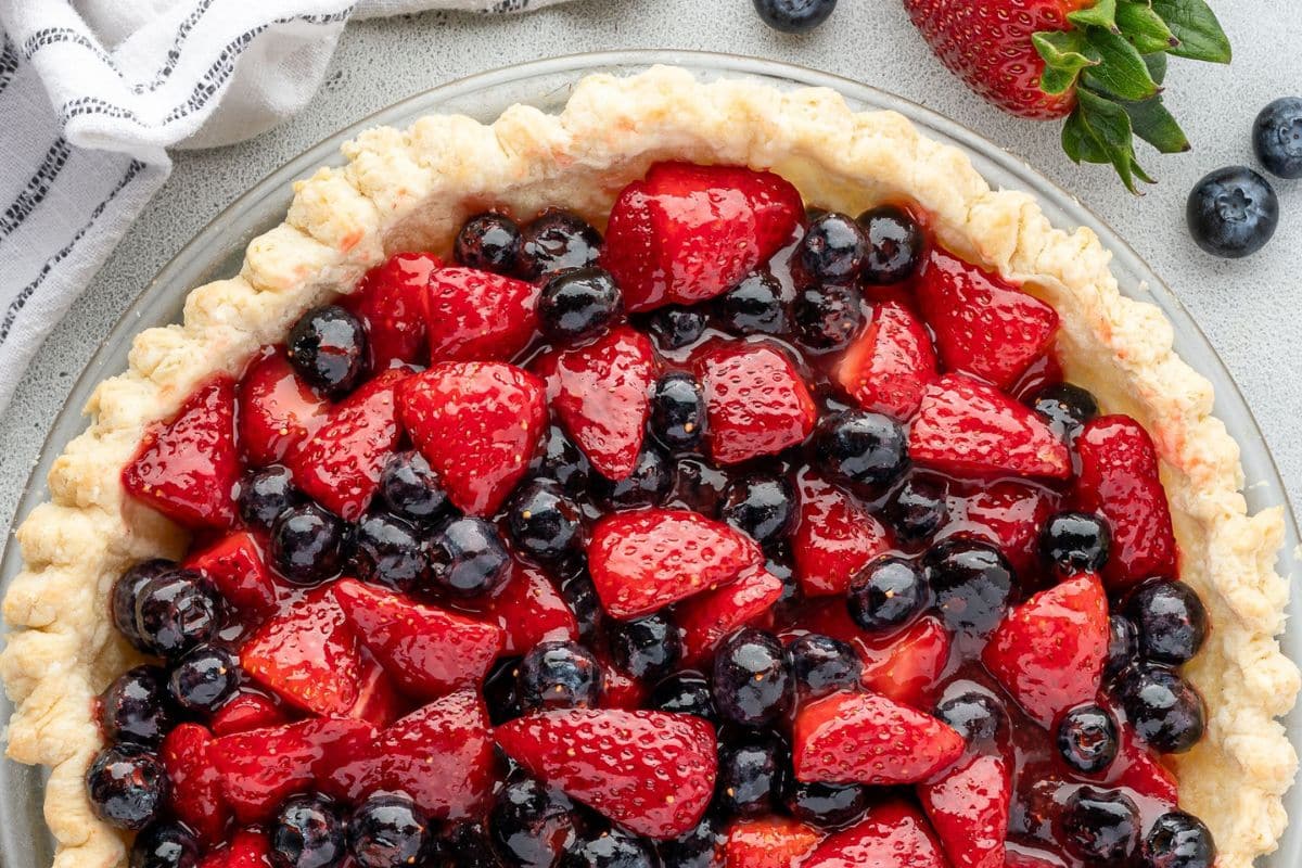 Close-up of a fruit pie topped with fresh strawberries and blueberries on a light surface.