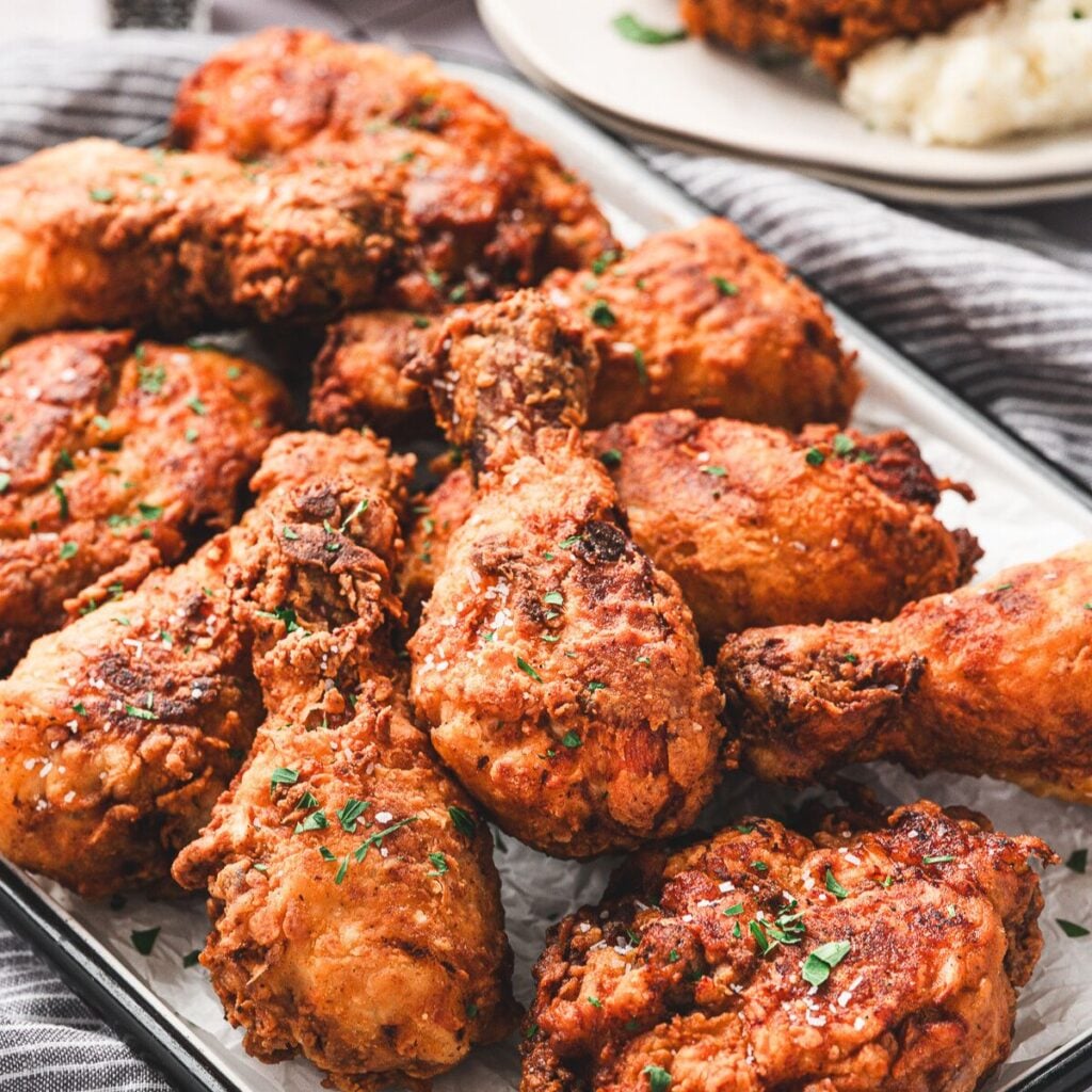 A tray of crispy fried chicken pieces garnished with herbs, with mashed potatoes visible in the background.