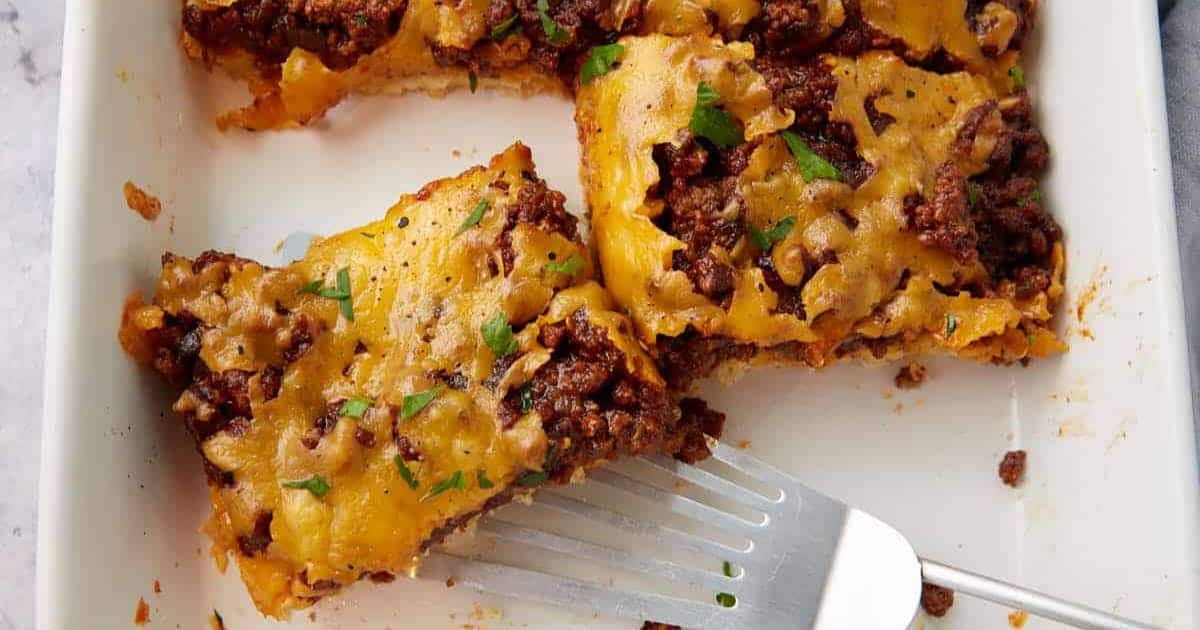 A spatula lifts a cheesy ground beef casserole from a white baking dish.