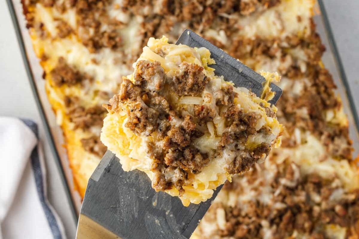 A spatula lifts a cheesy hash brown and sausage breakfast casserole from a glass baking dish.