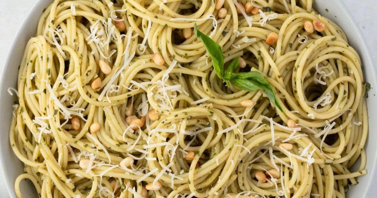 Spaghetti with pesto sauce, grated cheese, pine nuts, and a fresh basil garnish in a white bowl.