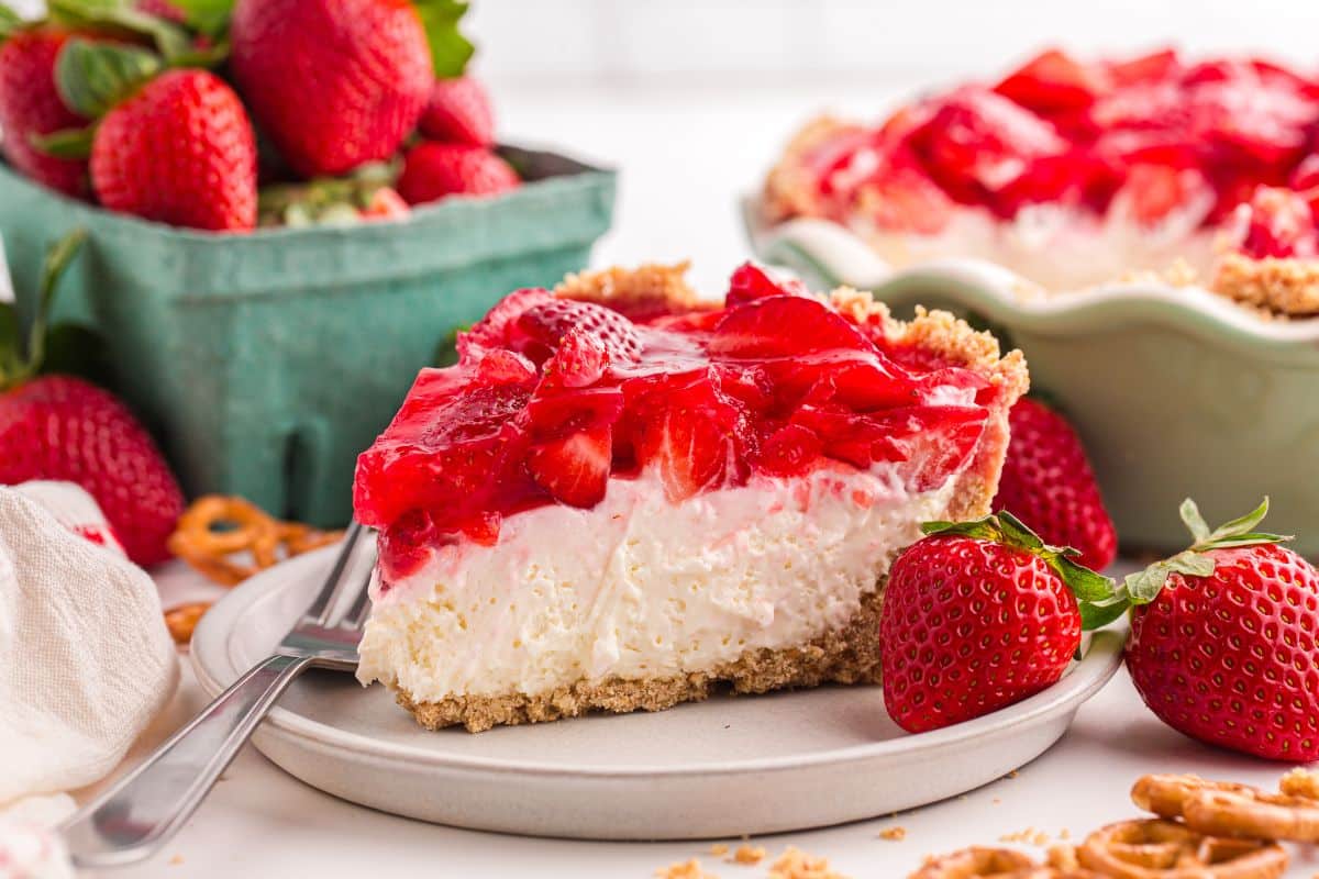 Slice of strawberry cheesecake on a plate, with fresh strawberries and a pie dish in the background.