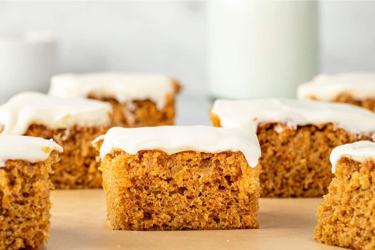 Squares of carrot cake with white frosting on top, arranged on parchment paper with a blurred background.
