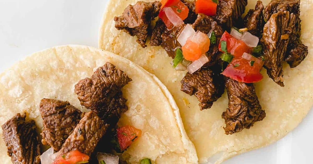 Two corn tortillas topped with grilled steak pieces and fresh chopped tomato salsa on a white plate.