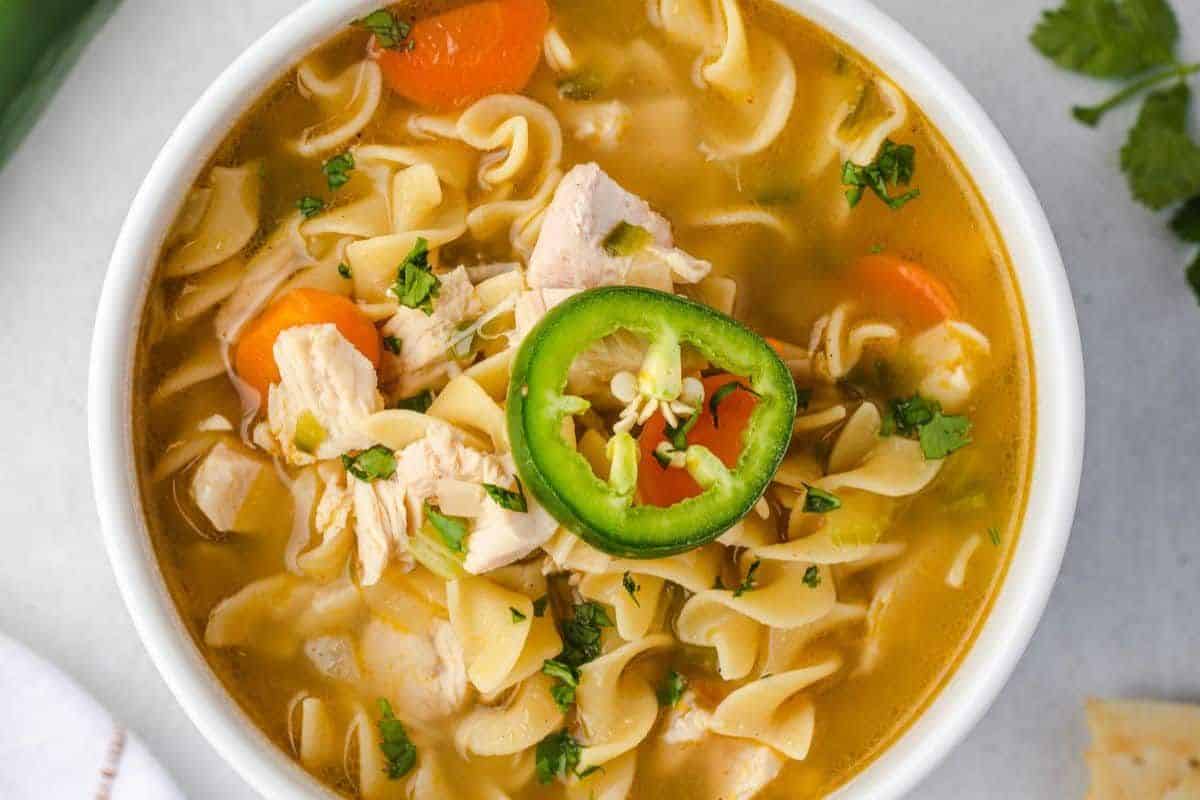 A bowl of chicken noodle soup with carrots, herbs, and a sliced jalapeƱo on top.