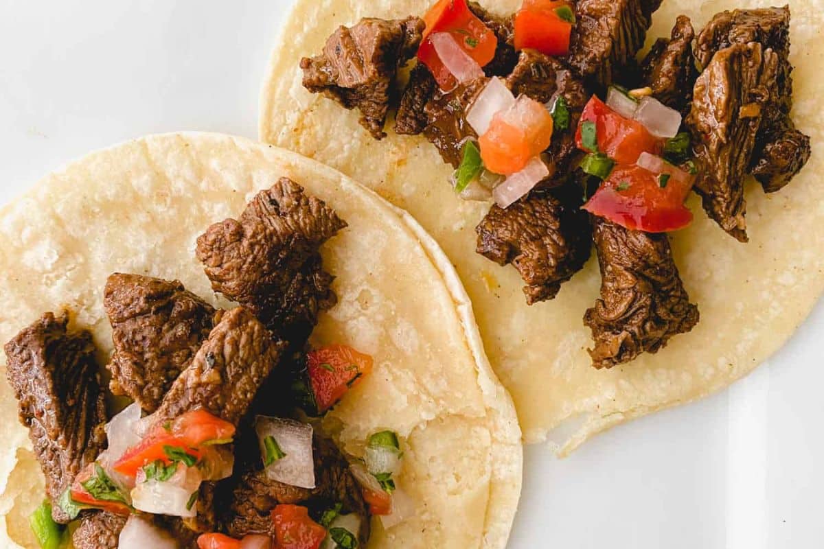 Two corn tortillas topped with grilled beef pieces and fresh pico de gallo on a white background.